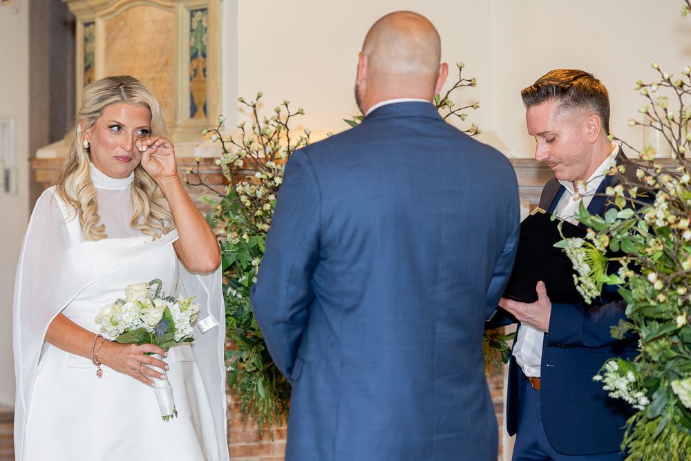 Bride wiping tears during emotional wedding ceremony while groom reads vows, with lush greenery backdrop.