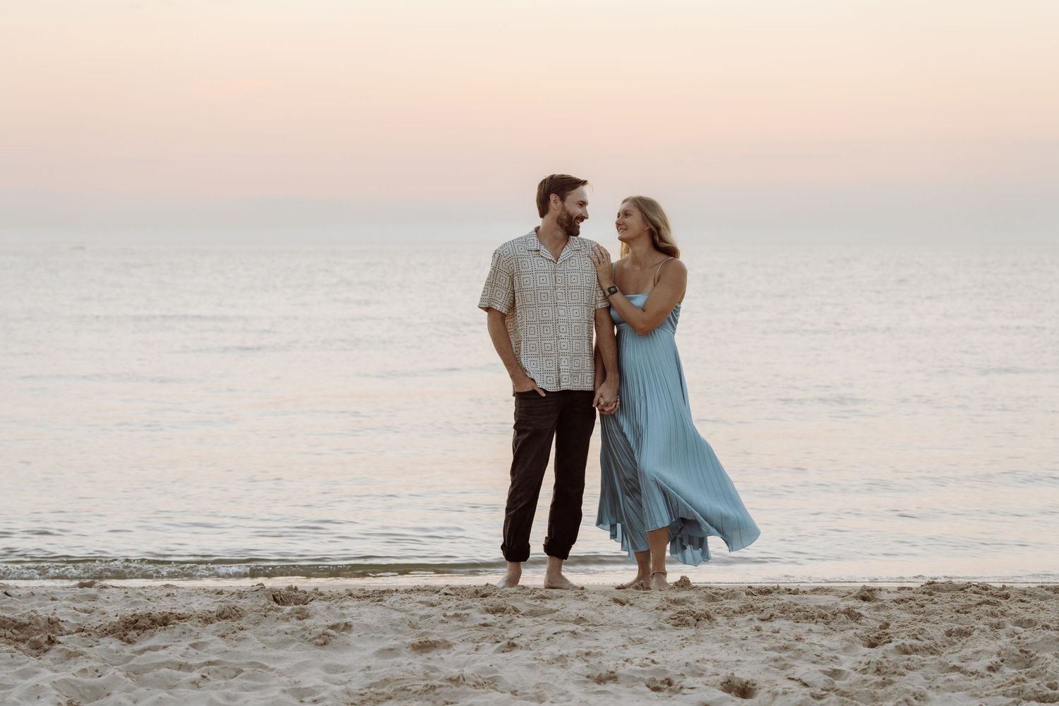 A couple stands together on a beach at sunset with the woman wearing a flowing light blue dress.