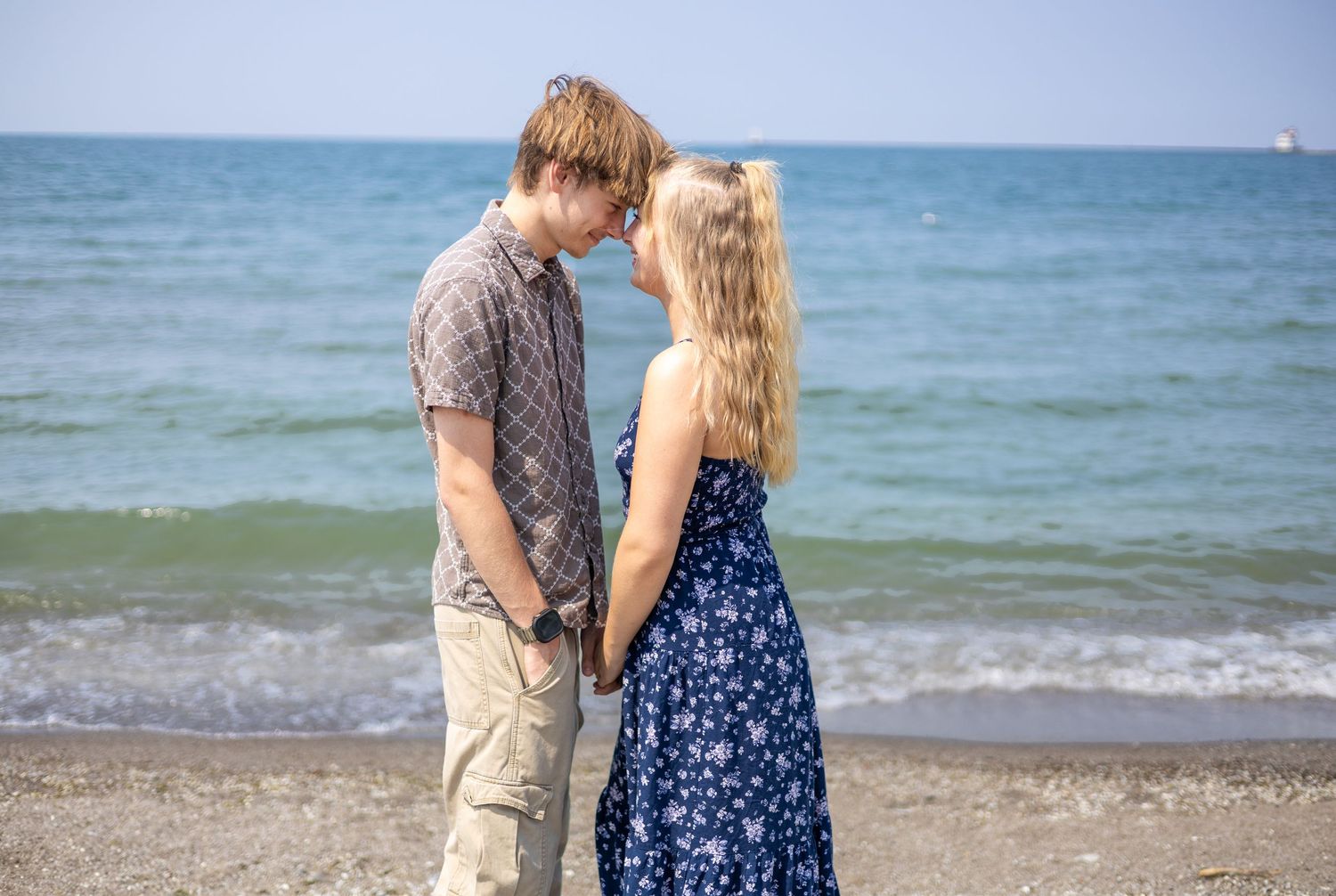 Young couple holding hands and facing each other romantically on a sunny beach by the ocean.