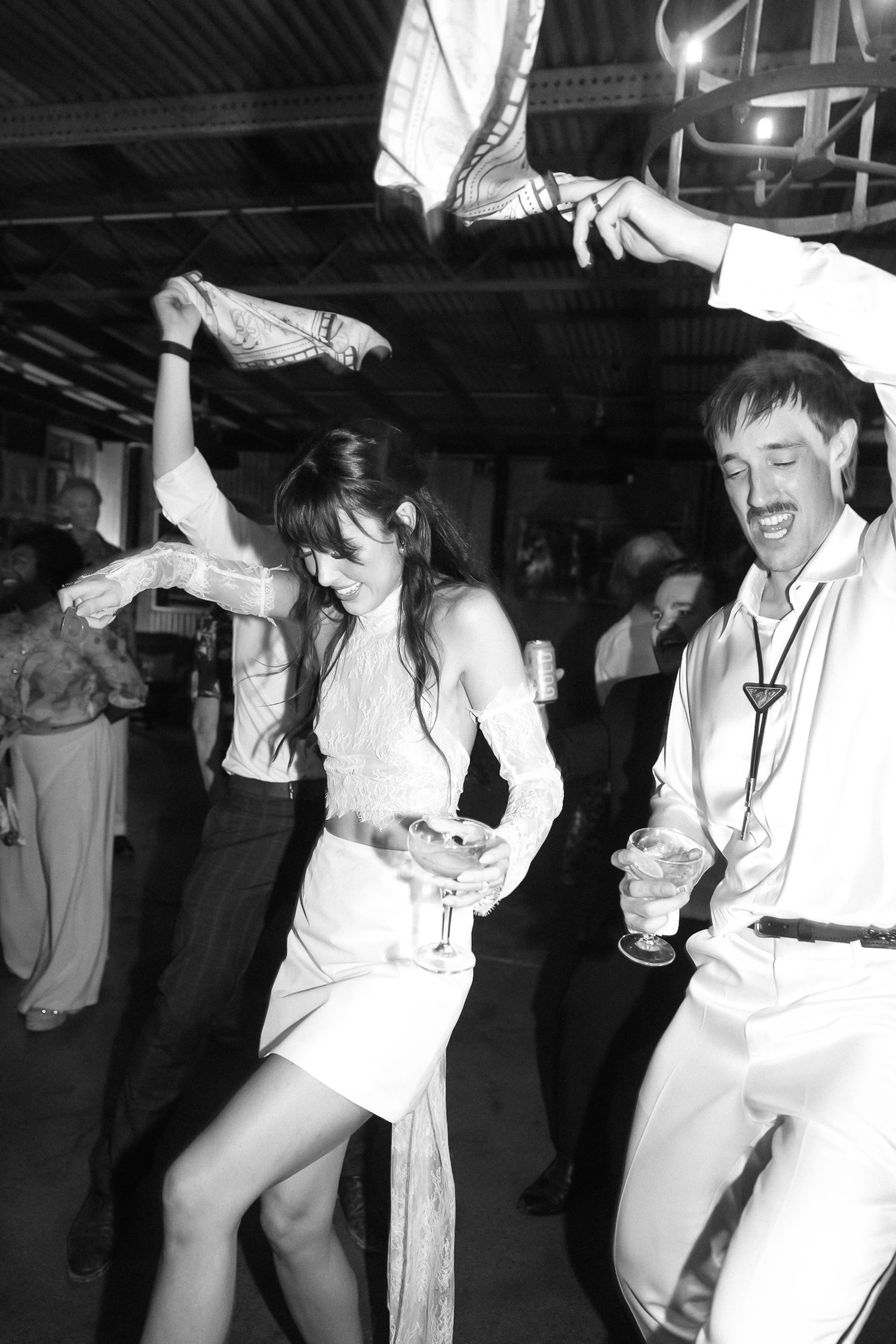 Black and white photo of a couple dancing energetically at a party, holding drinks and raising arms in celebration.