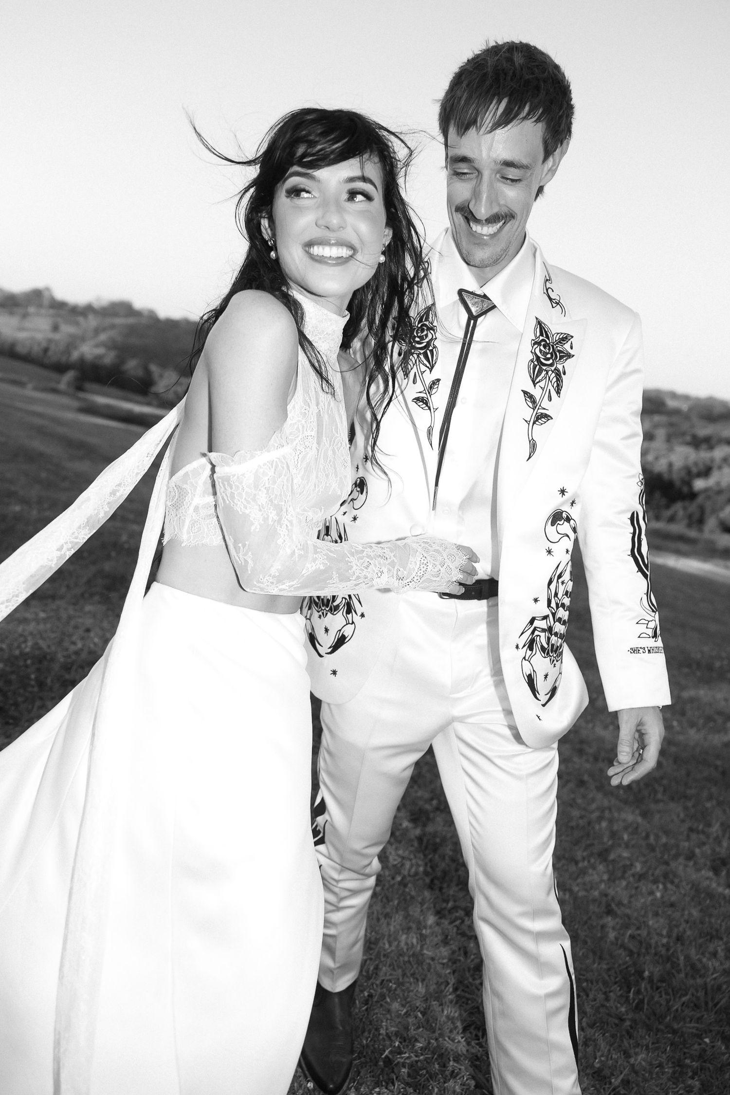 Black and white photo of a smiling bride and groom in ornate white outfits walking outdoors on their wedding day.