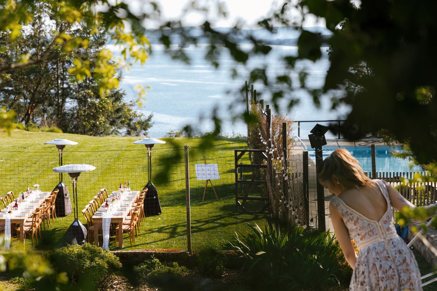 Outdoor wedding reception setup on lush green lawn overlooking ocean, with long white tables and chairs arranged elegantly.