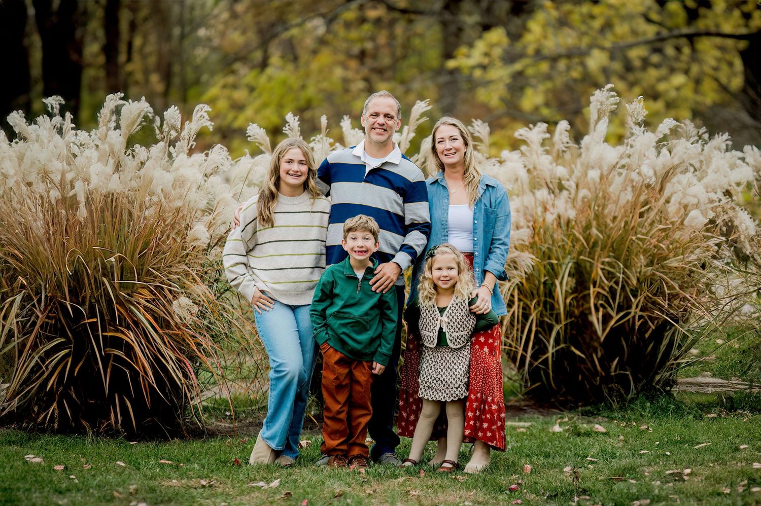 Family of five smiling outdoors in fall, posing in front of tall ornamental grasses in a lush green park.