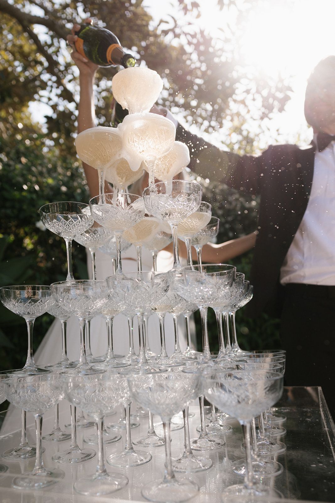 Elegant champagne tower with glasses being filled at a sunlit outdoor wedding reception.