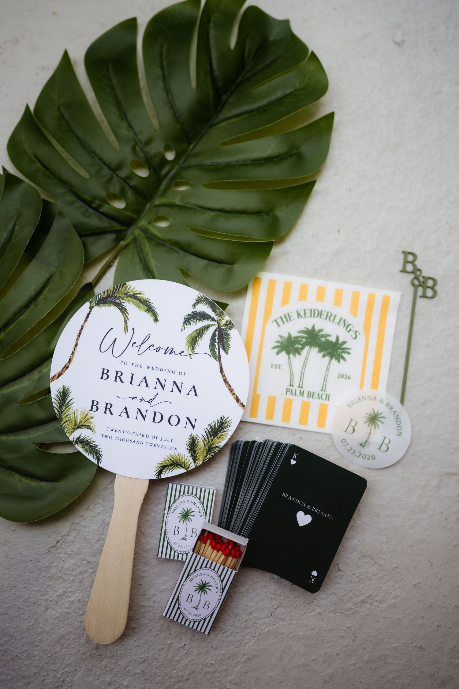 Tropical wedding welcome fan, palm tree matchbooks, playing cards, and striped stationery suite with monstera leaf backdrop.