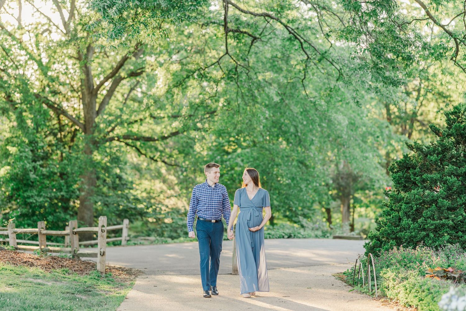 A couple walks hand-in-hand on a sunlit garden path surrounded by lush green trees during a maternity photoshoot.