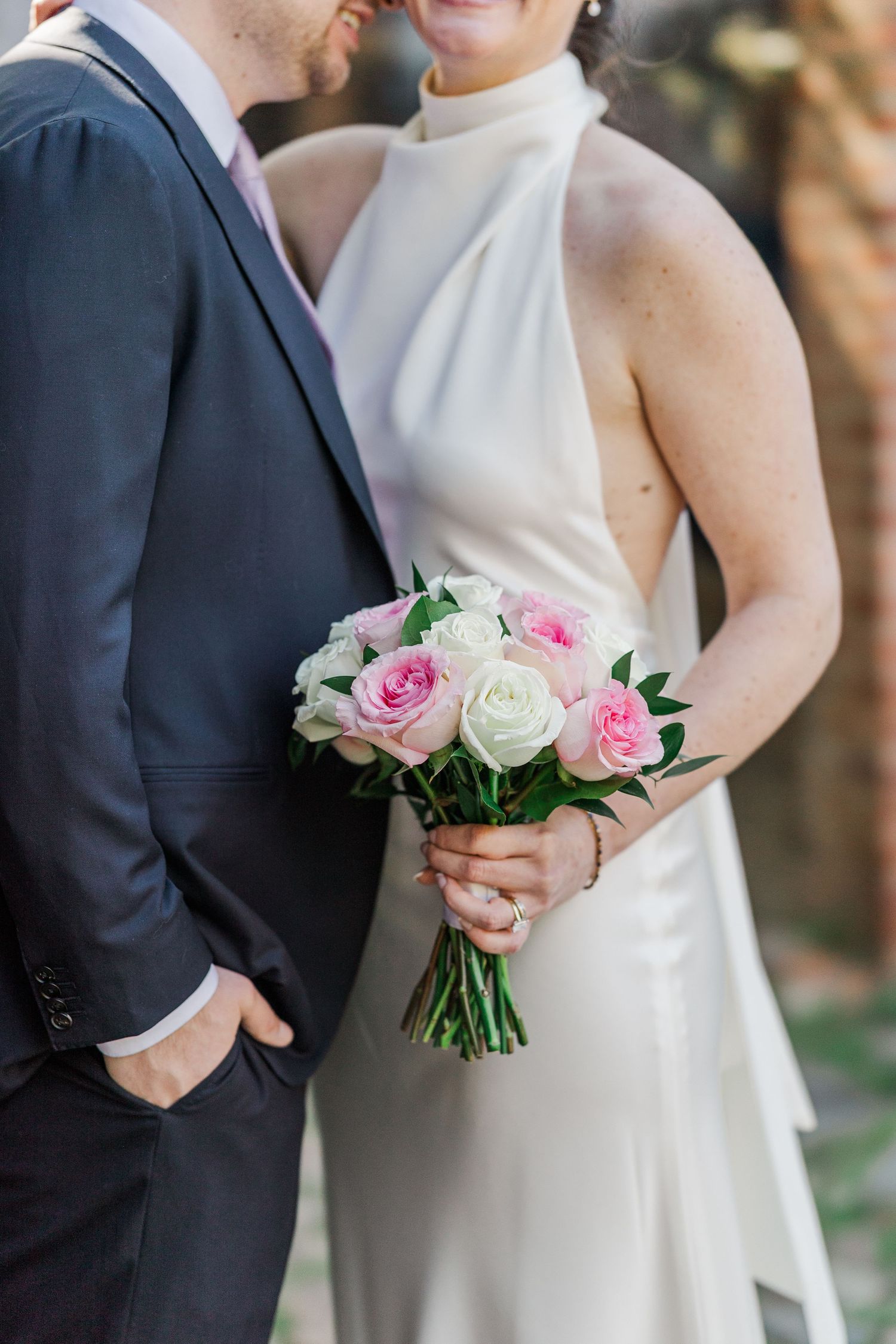 Bride in white halter gown holds pink and white rose bouquet while embracing groom in navy suit outdoors.