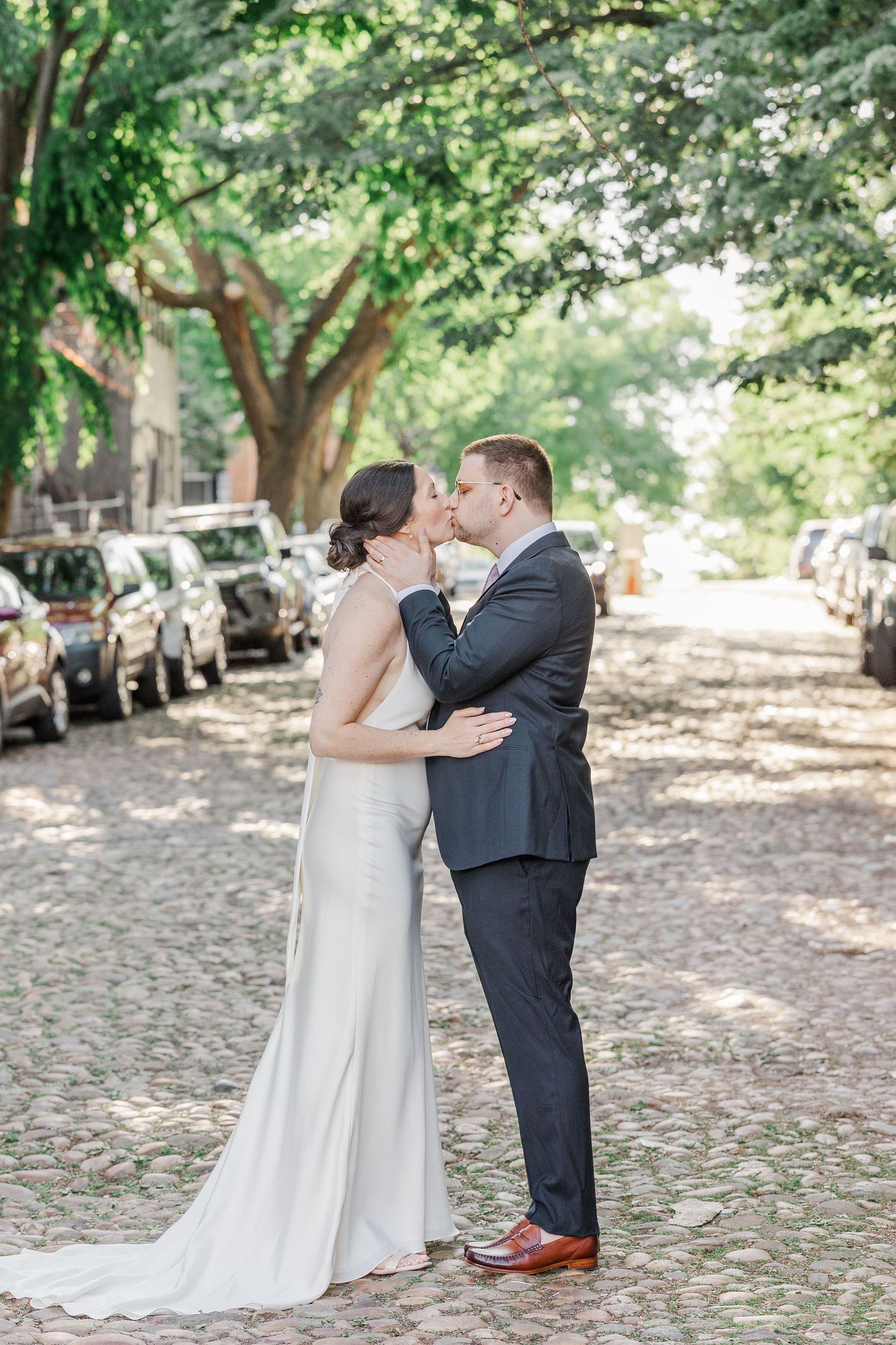 Bride and groom share a kiss on a cobblestone street lined with parked cars and lush green trees.