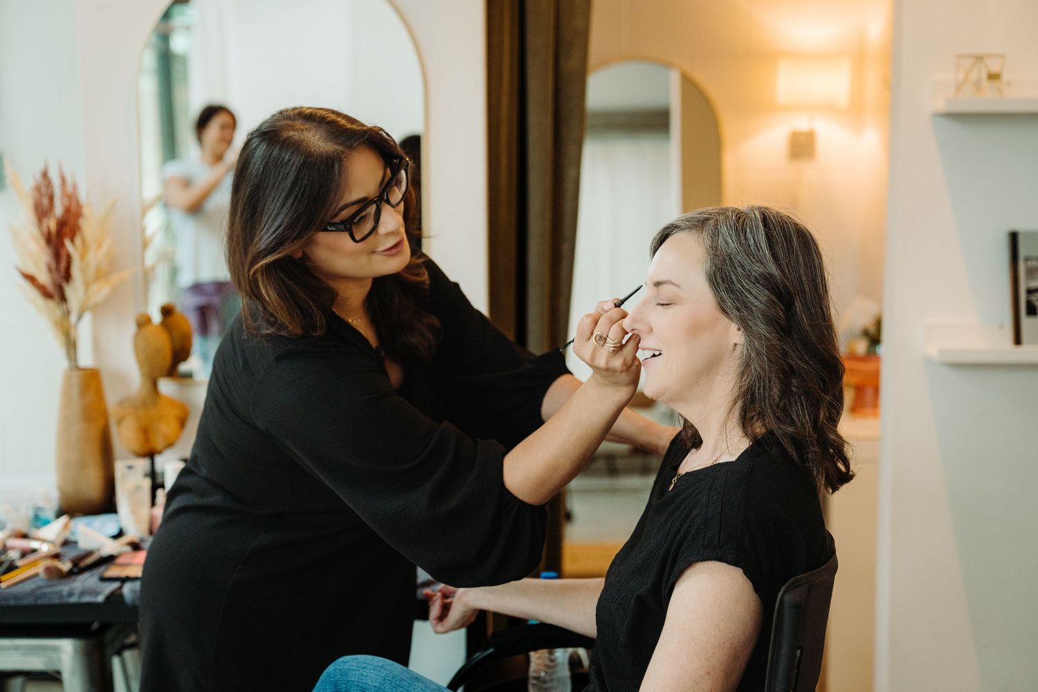 A professional makeup artist applying eye makeup to a seated woman in a bright salon studio.