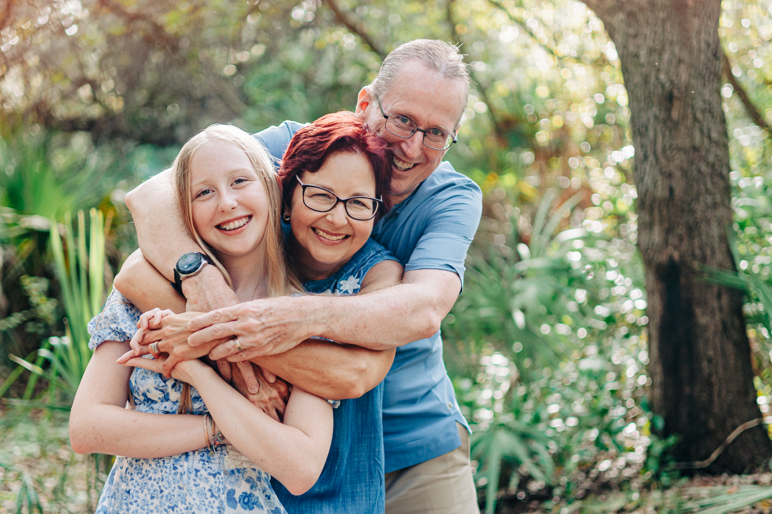 Happy family of three hugging outdoors in a lush green garden, smiling joyfully together at Boynton beach park.