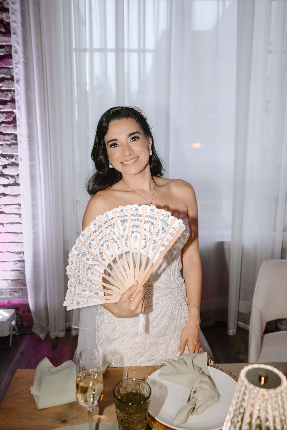 Smiling bride in white off-shoulder gown holding decorative lace fan at elegantly set wedding reception table.