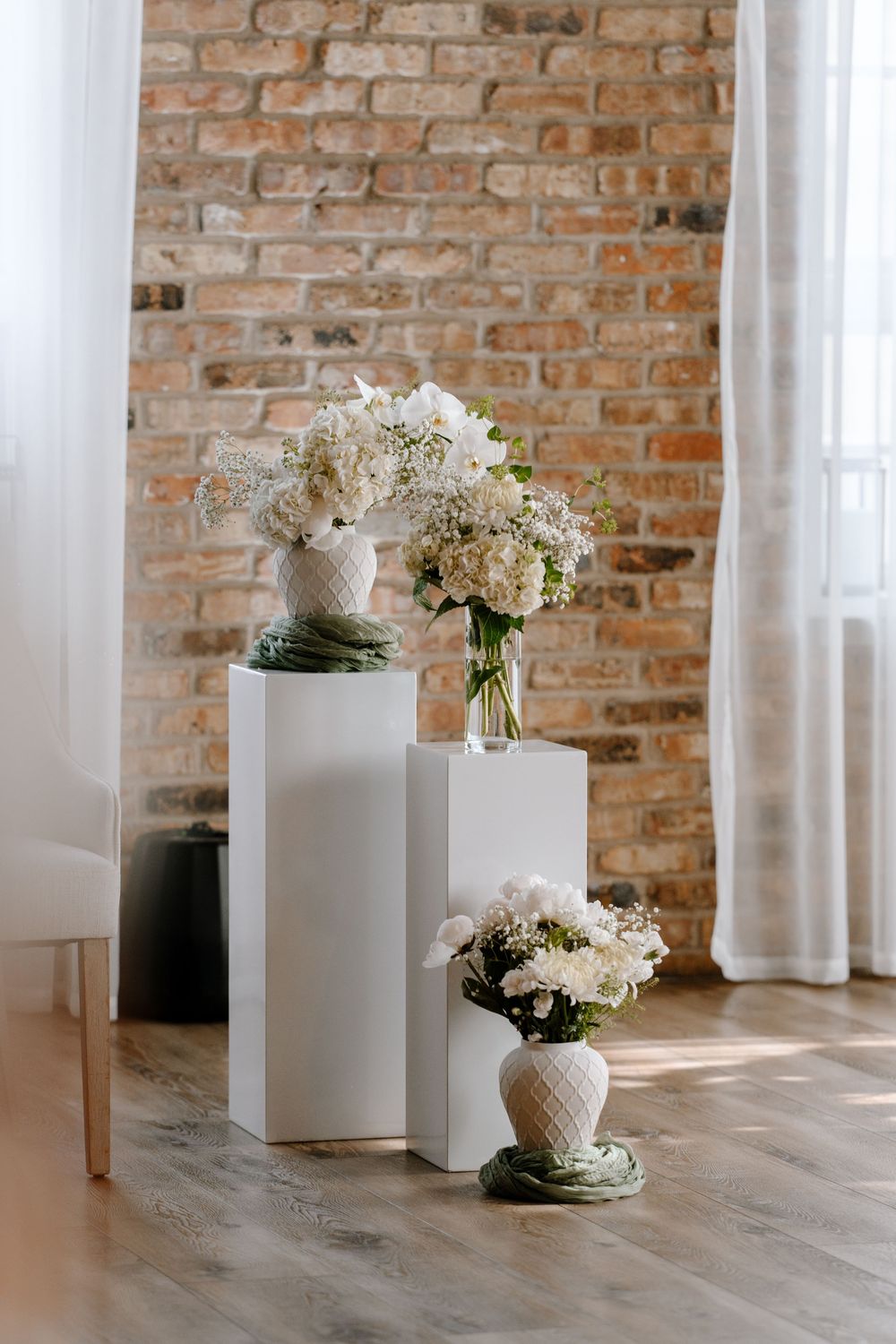 White floral arrangements in vases displayed on white pedestals against a rustic brick wall with sheer curtains.