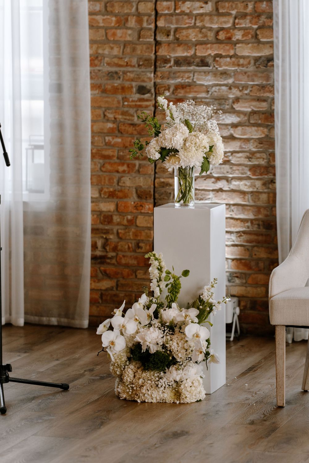 White floral arrangements on tiered white pedestals against rustic brick wall with hardwood floors in elegant wedding decor.
