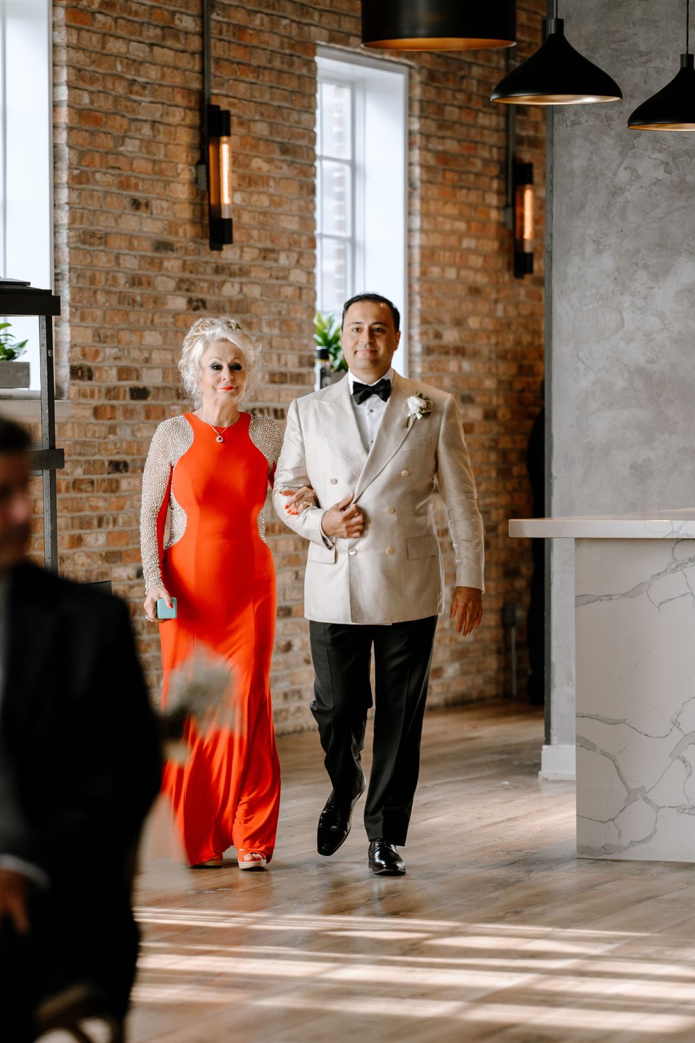 Elegant couple walking arm-in-arm at wedding venue, woman in orange gown and man in ivory tuxedo with bow tie.