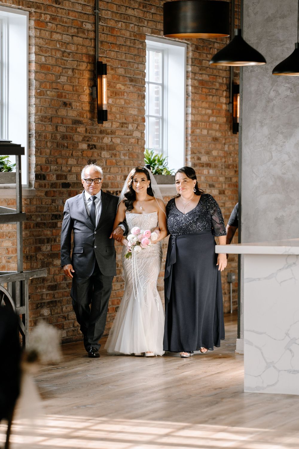Bride in white gown walks down aisle with parents in elegant brick-walled venue holding pink bouquet.