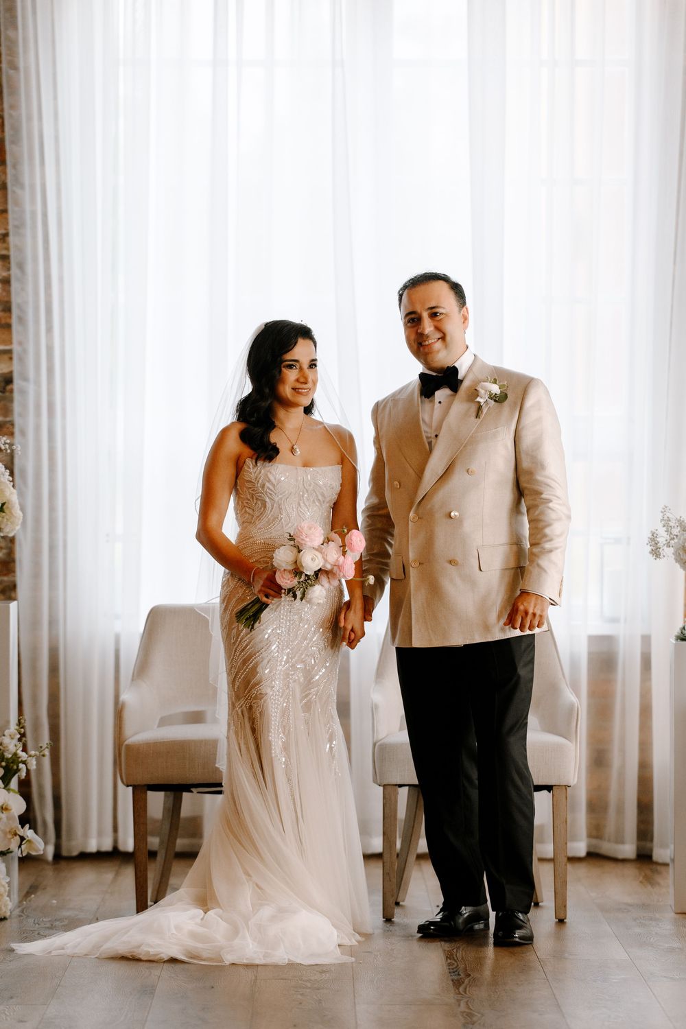 Bride in beaded strapless gown holding pink bouquet stands with groom in ivory double-breasted tuxedo at wedding ceremony.