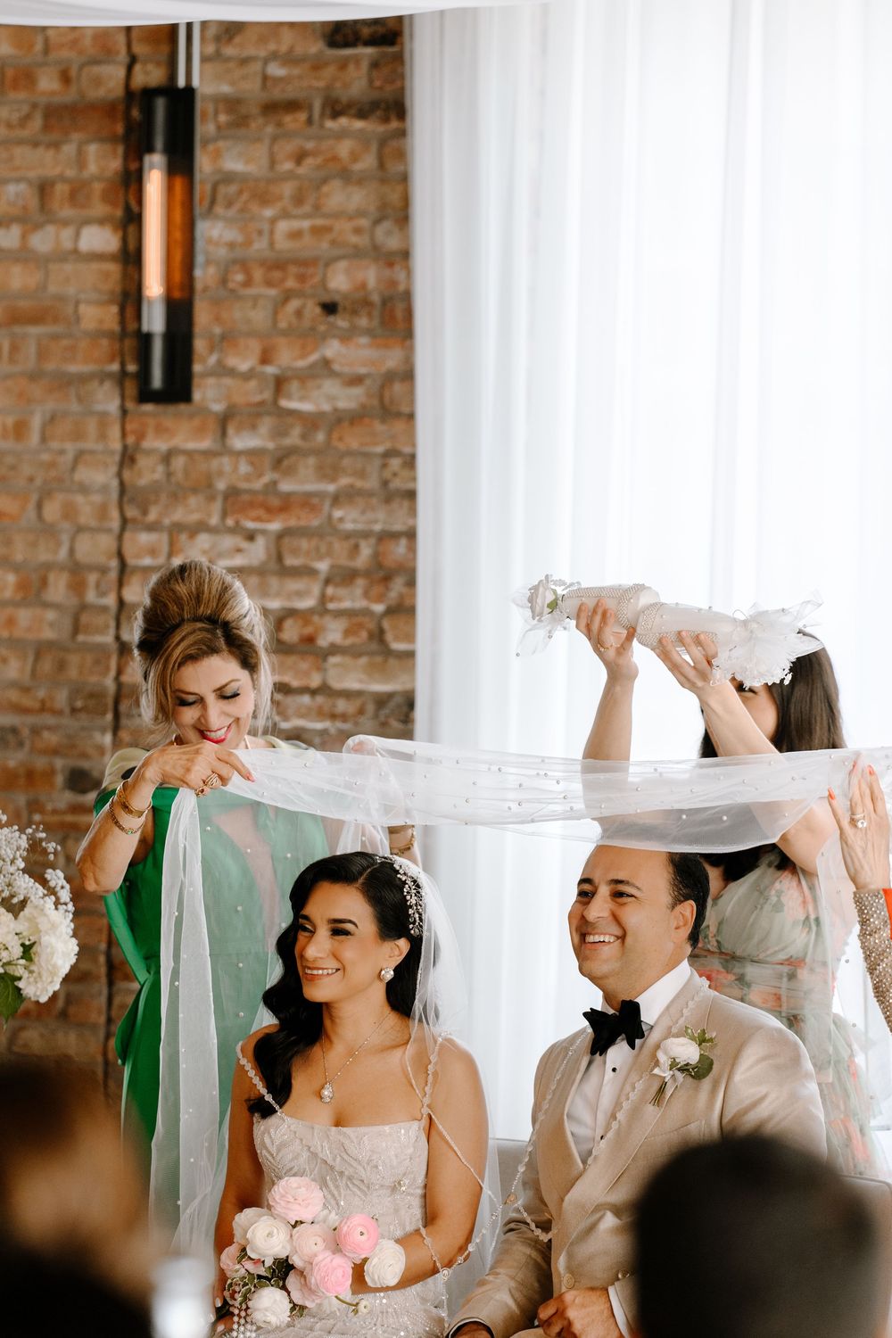 Bride and groom seated during wedding ceremony while bridesmaids hold a sheer veil overhead in an elegant venue.