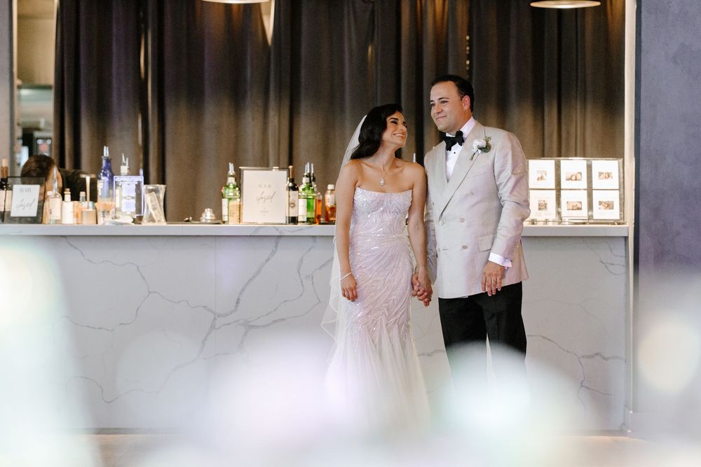 Bride in strapless lace gown and groom in white tuxedo jacket hold hands near marble bar at elegant wedding reception.