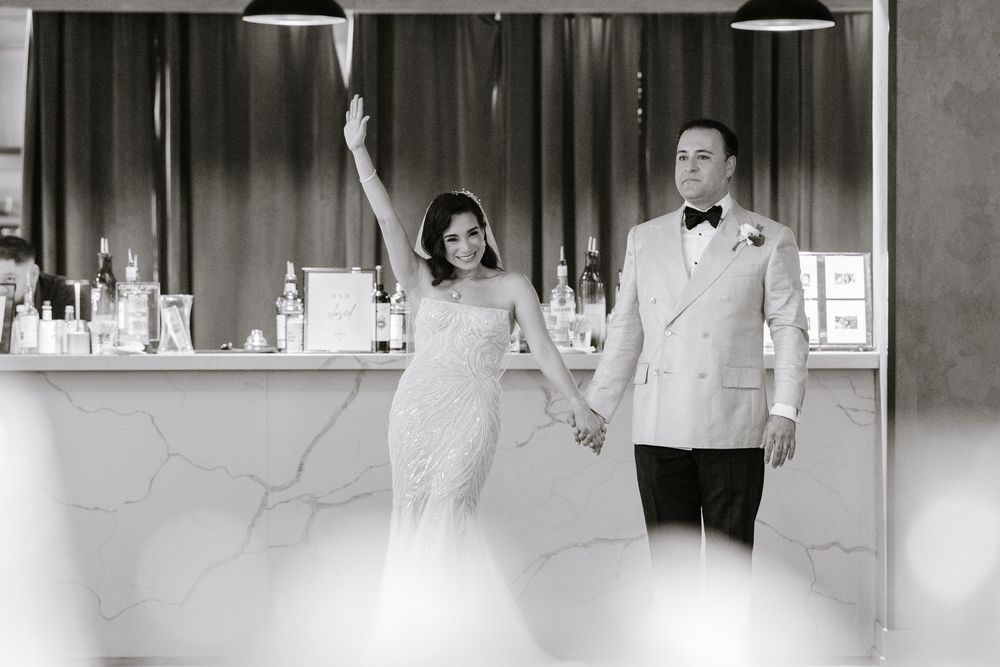 Black and white photo of bride and groom holding hands at wedding reception bar, bride raising arm in celebration.
