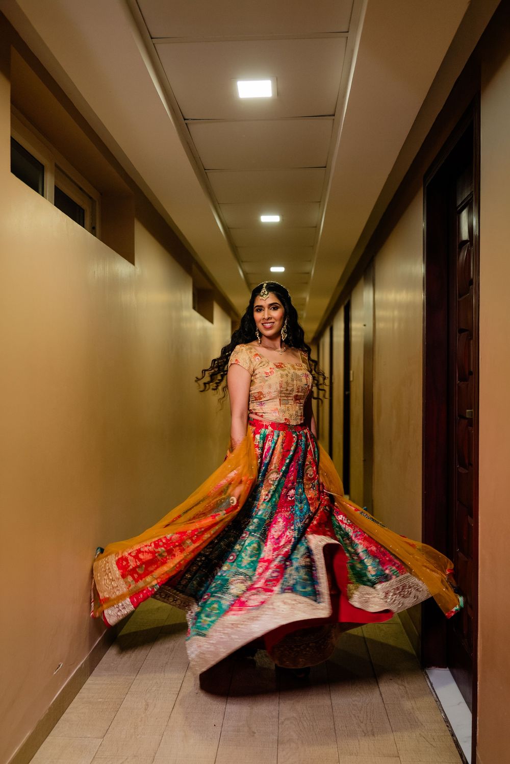 A bride twirls in a vibrant multicolor lehenga with gold dupatta in a hotel corridor.