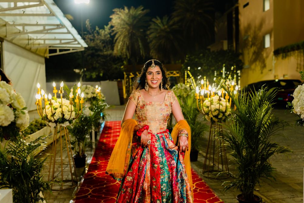 Bride in vibrant red and green lehenga walks down floral-lit aisle at elegant Indian wedding ceremony.