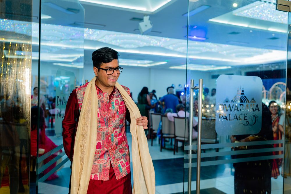 Man in traditional Indian attire with red kurta, cream shawl, and glasses posing at a decorated venue.