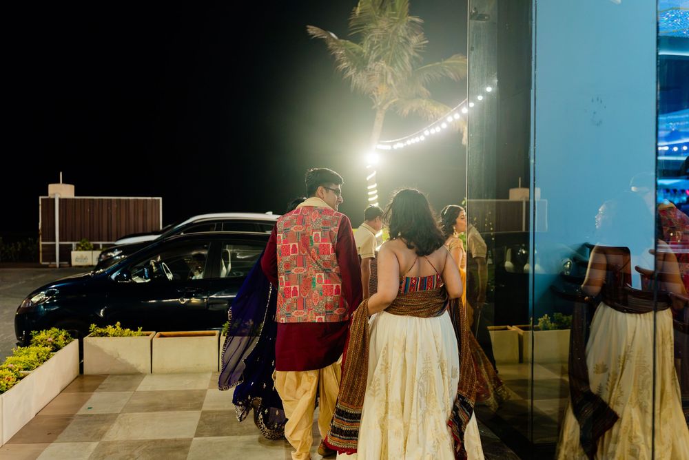 Couple in traditional Indian attire walking outdoors at night near a decorated venue with string lights.
