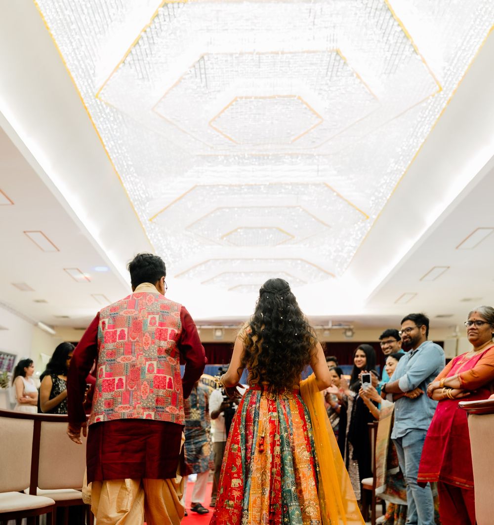 Indian couple in traditional attire walking into elegant wedding venue with ornate crystal ceiling.