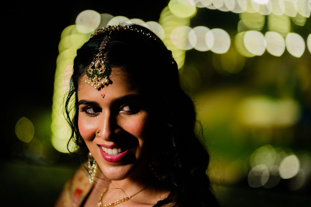 Indian bride smiling at night wearing traditional gold maang tikka jewelry with bokeh lights in background.