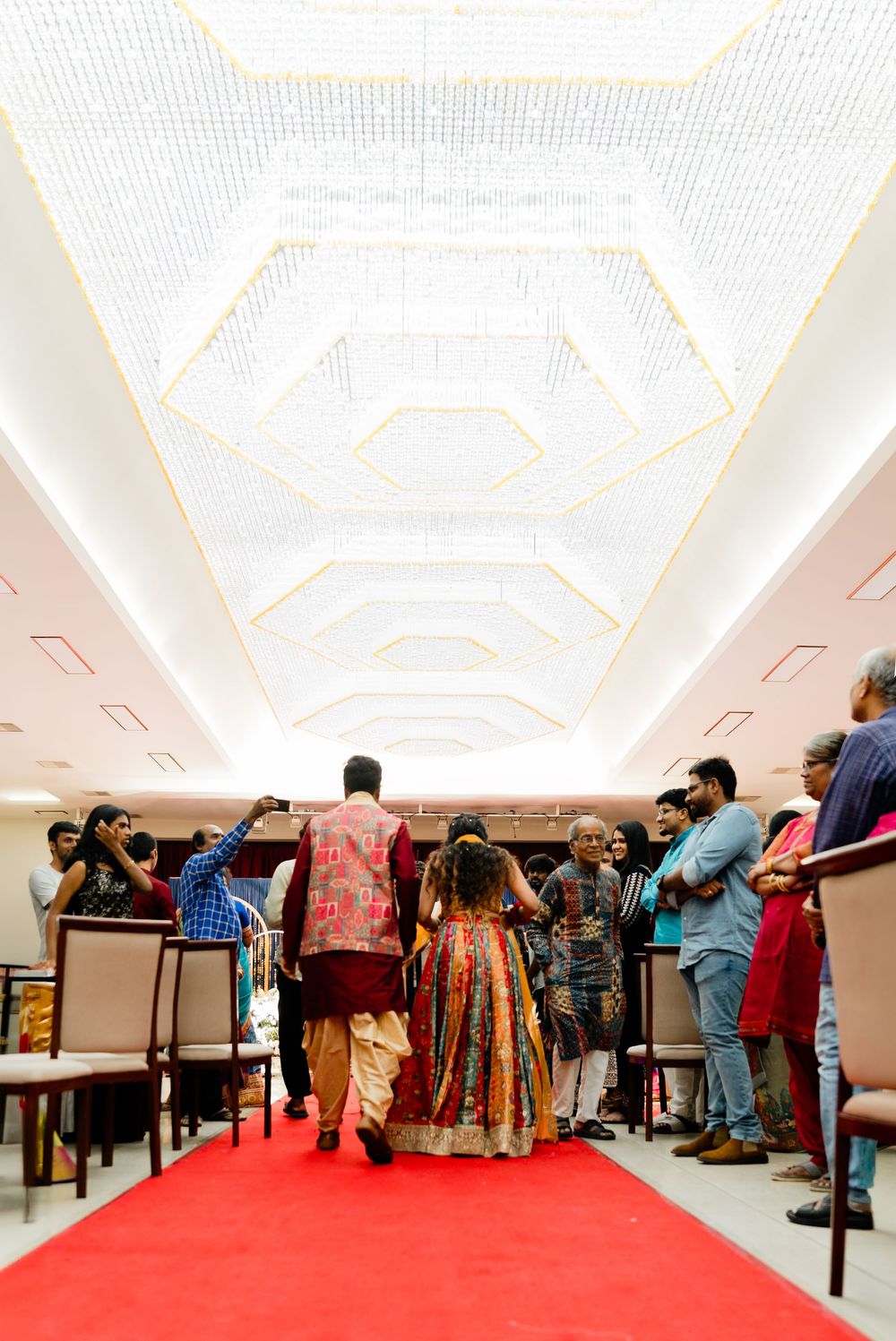Indian wedding ceremony guests walk down red carpet aisle under ornate geometric ceiling in elegant venue.