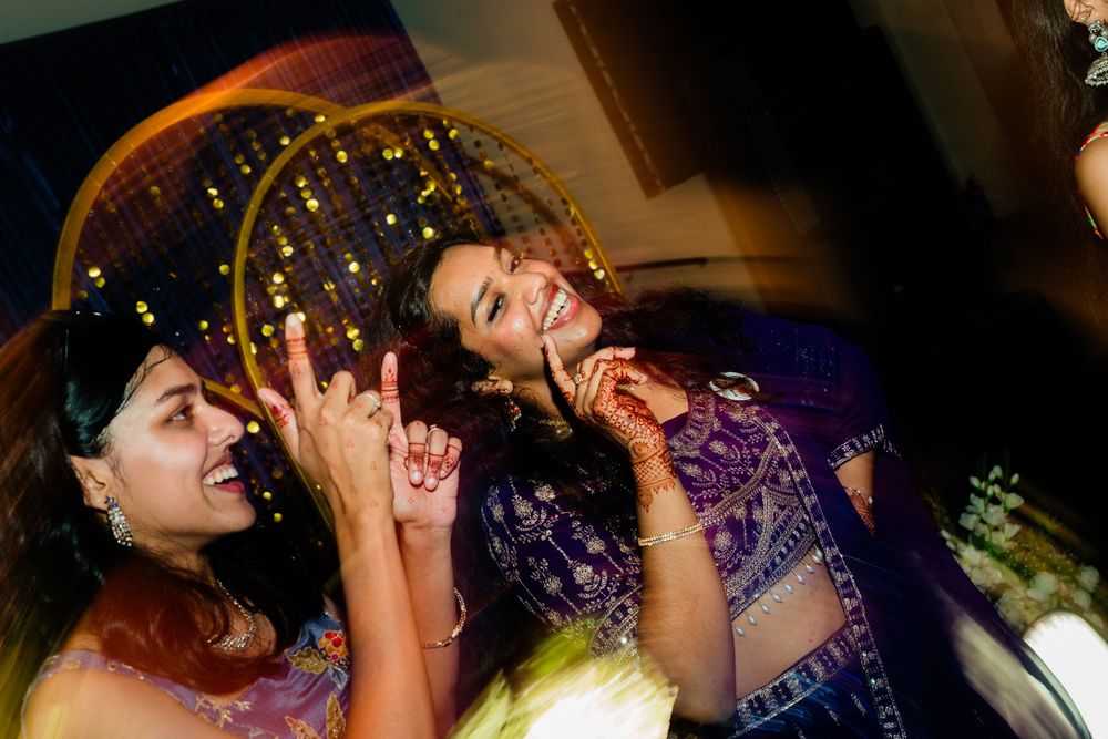 Two women in purple and blue Indian attire laughing and dancing at a festive event.