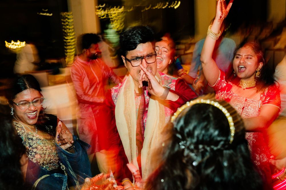 Groom in red sherwani sings into a microphone surrounded by joyful guests at a lively Indian wedding party.