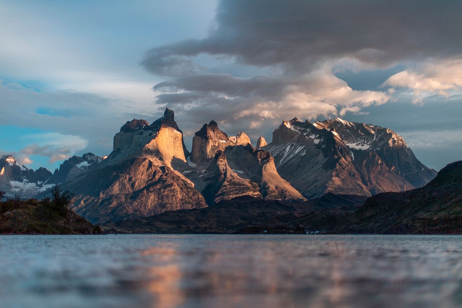 Dramatic mountain peaks of Torres del Paine reflect in calm Patagonian lake waters at golden sunset under stormy skies.