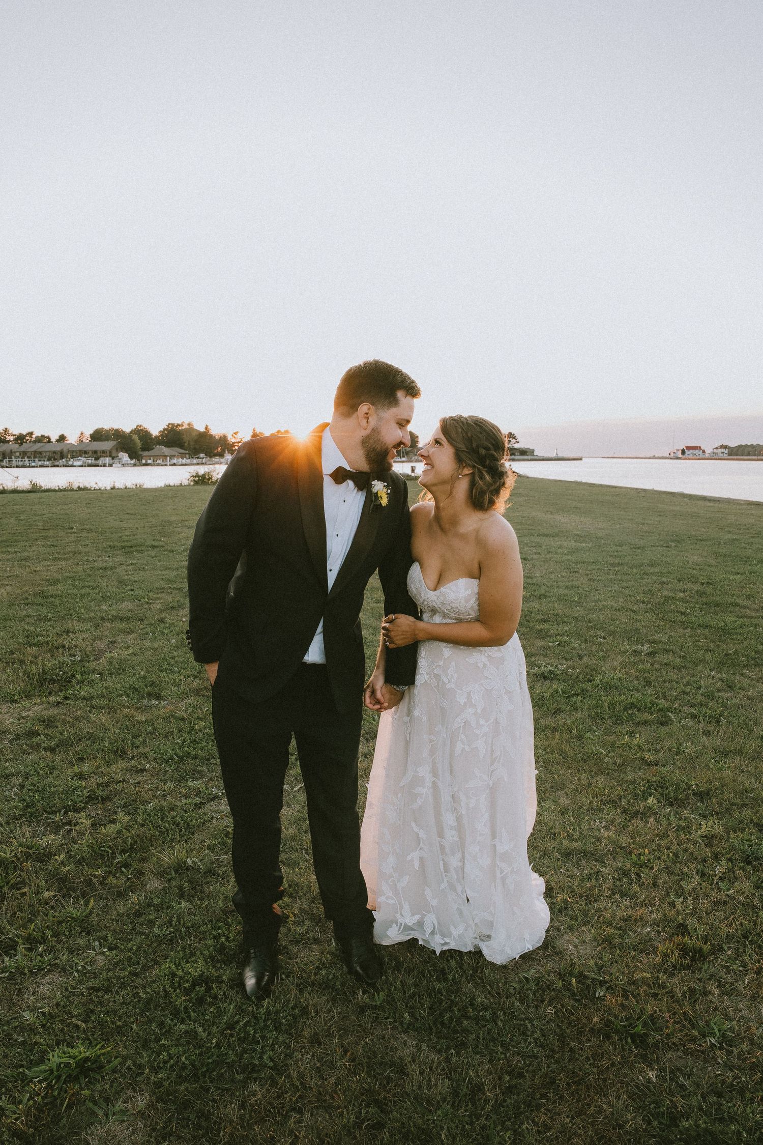 Bride and groom share a romantic moment at sunset by the waterfront, dressed in wedding attire on a green lawn.