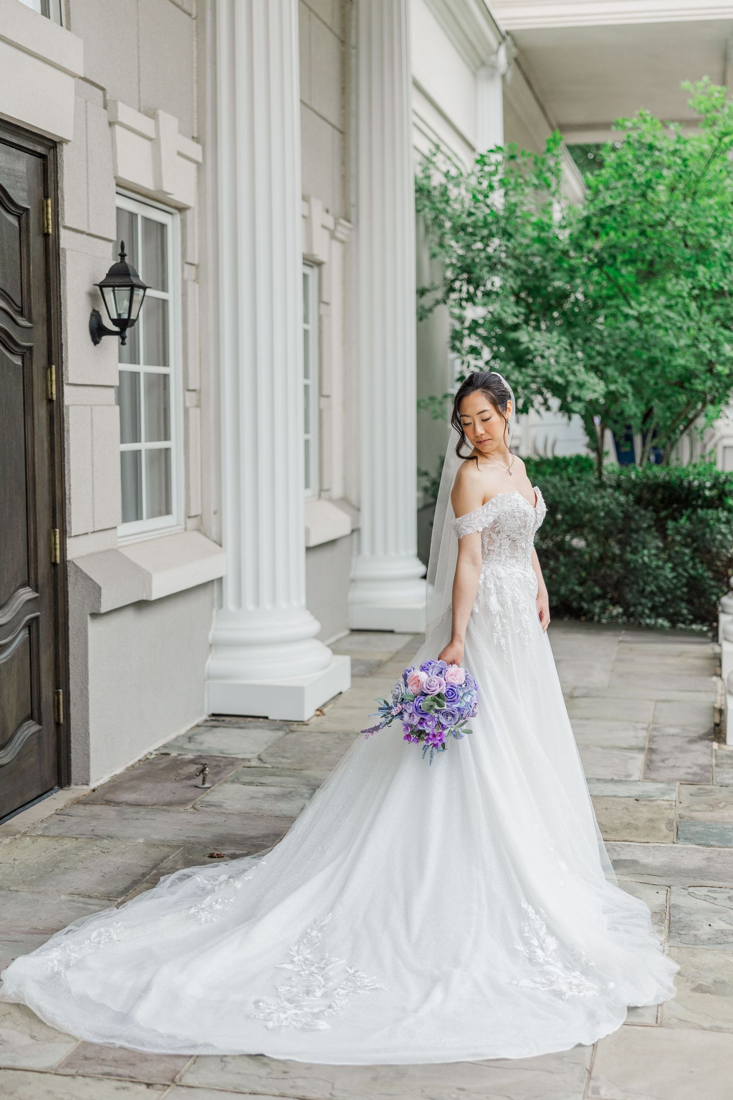 Bride in off-shoulder white lace gown with long train holds purple bouquet beside elegant white columns.