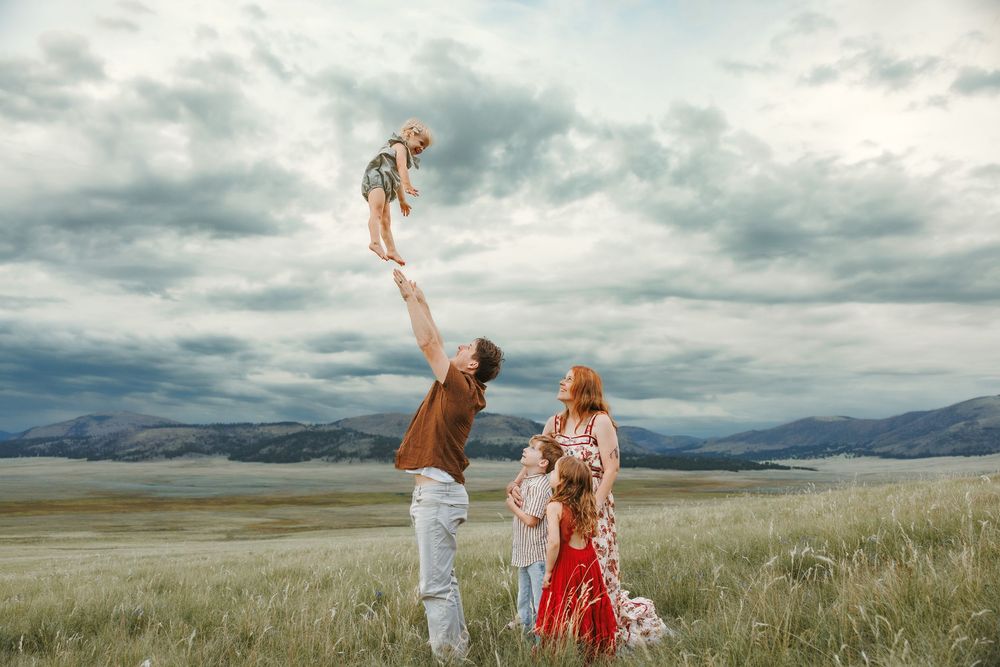 Family enjoying outdoor photoshoot in grassy meadow, father lifting child overhead, mountains and cloudy sky backdrop.