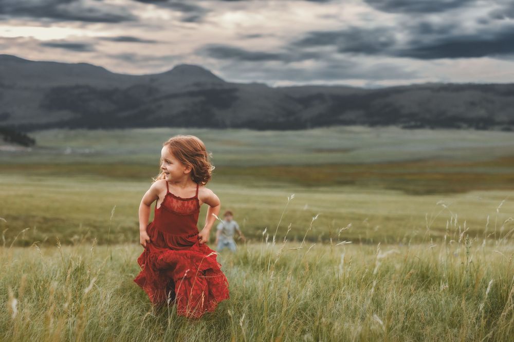 Young girl in a red dress running through a vast green meadow with dramatic mountain scenery in the background.