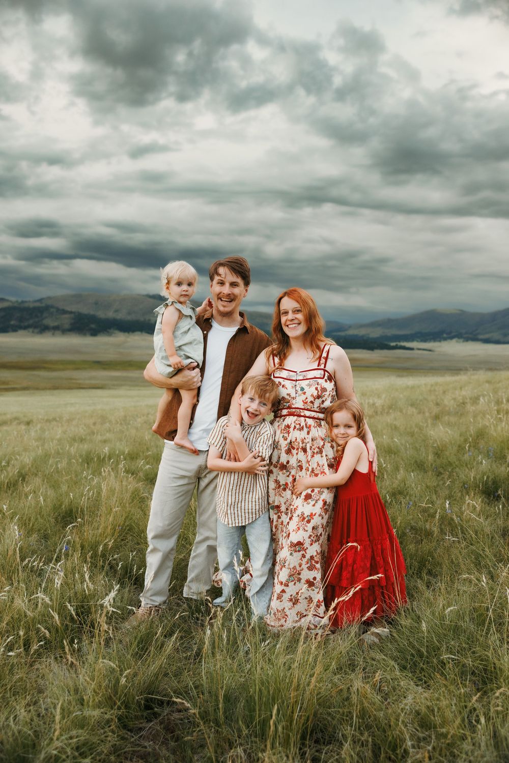 Family of five posing in a green meadow with mountains and dramatic cloudy sky in the background.