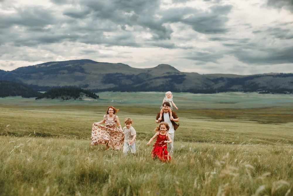 Family running joyfully through green meadow with mountain backdrop under dramatic cloudy sky.