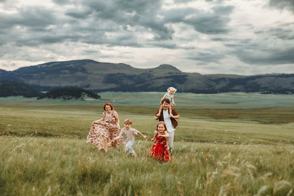 Family with children joyfully running through a green field with mountains and cloudy skies in the background.