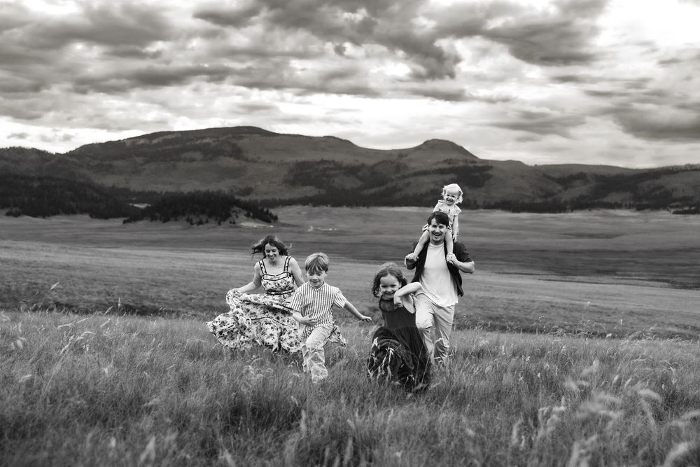 Black and white photo of a joyful family playing in a meadow with rolling hills and dramatic clouds behind them.