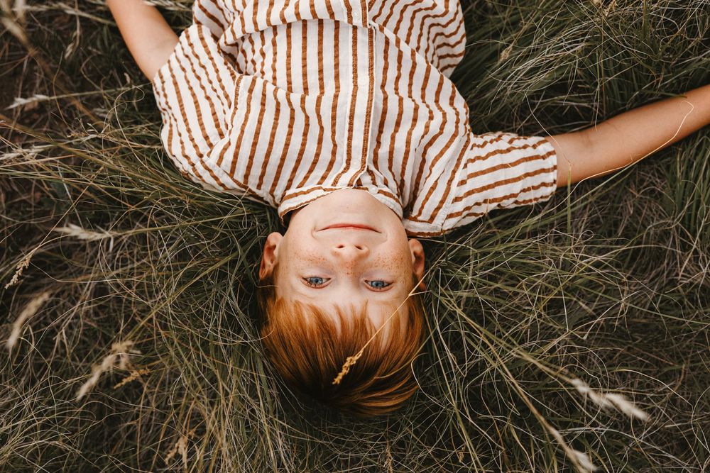 Child in a striped dress lying in tall grass, looking up at the camera with a peaceful expression.