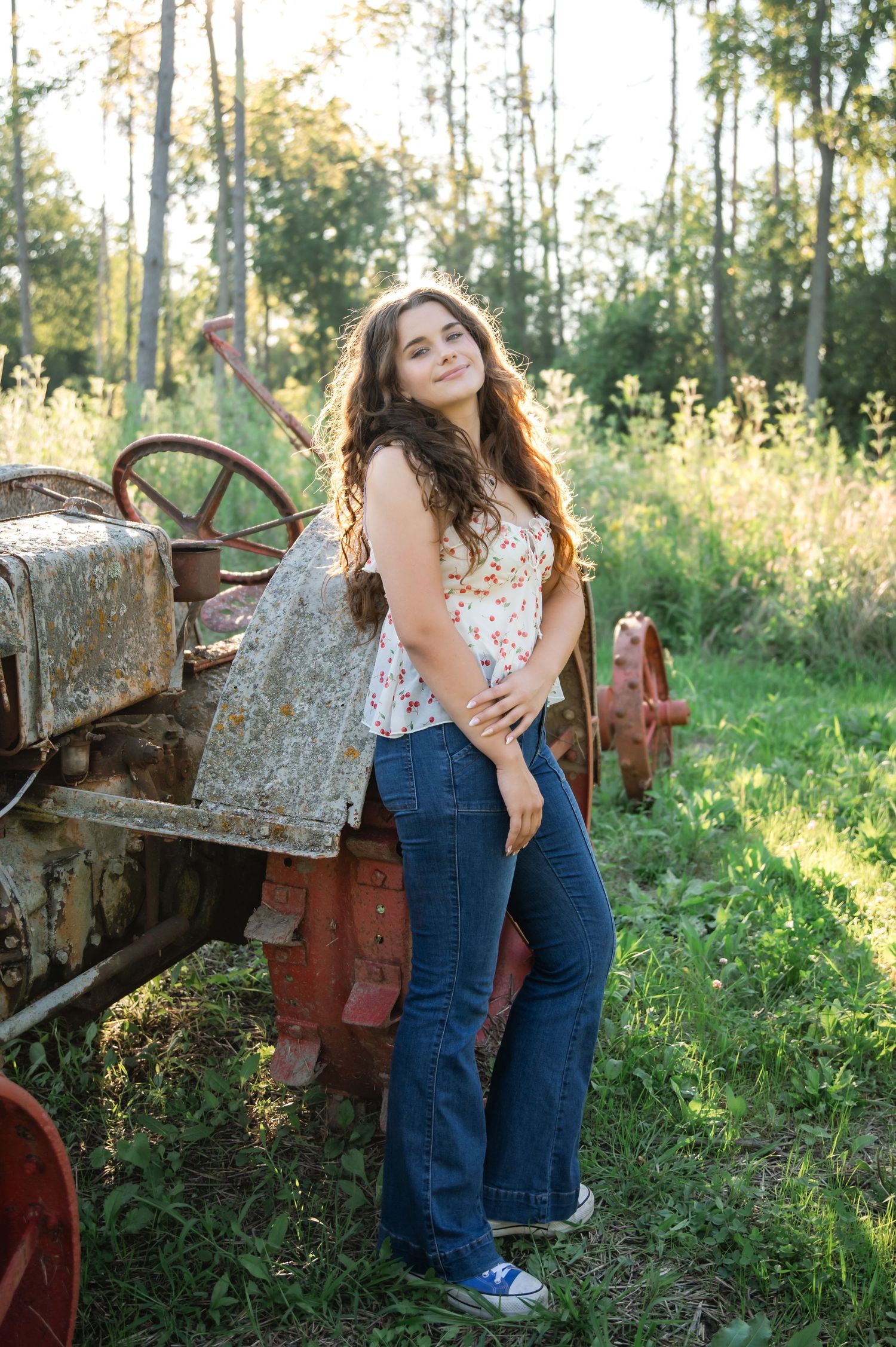 Young woman in floral top and flared jeans leaning against a rustic vintage tractor in a lush green field.
