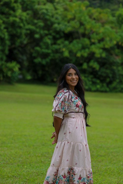 A smiling woman in a floral embroidered maxi dress poses in a lush green garden setting.