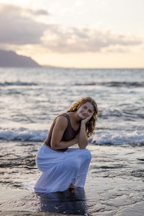 Young woman in white skirt crouching in ocean waves at sunset with mountain silhouette behind her.