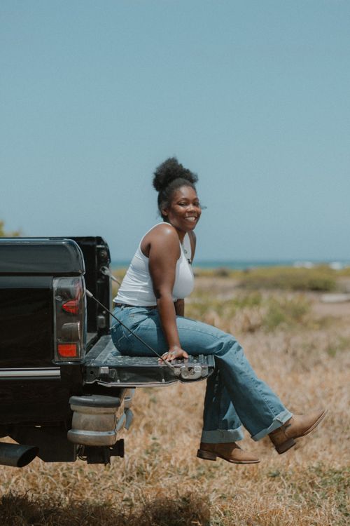 A smiling young woman sits on a truck tailgate in jeans and white tank top in a sunny field.