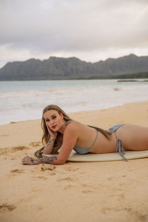 A tattooed blonde woman in a grey bikini lies on a surfboard on a sandy beach with mountains behind.
