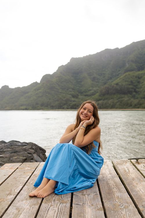 Young woman in blue dress sitting barefoot on wooden dock with green mountain and water backdrop.