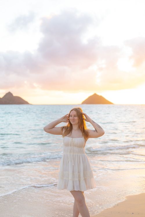 Young woman in white sundress standing on beach at sunrise with island silhouettes behind her.
