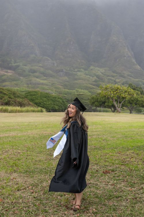 Smiling graduate in cap and gown posing outdoors with misty green mountains in the background.